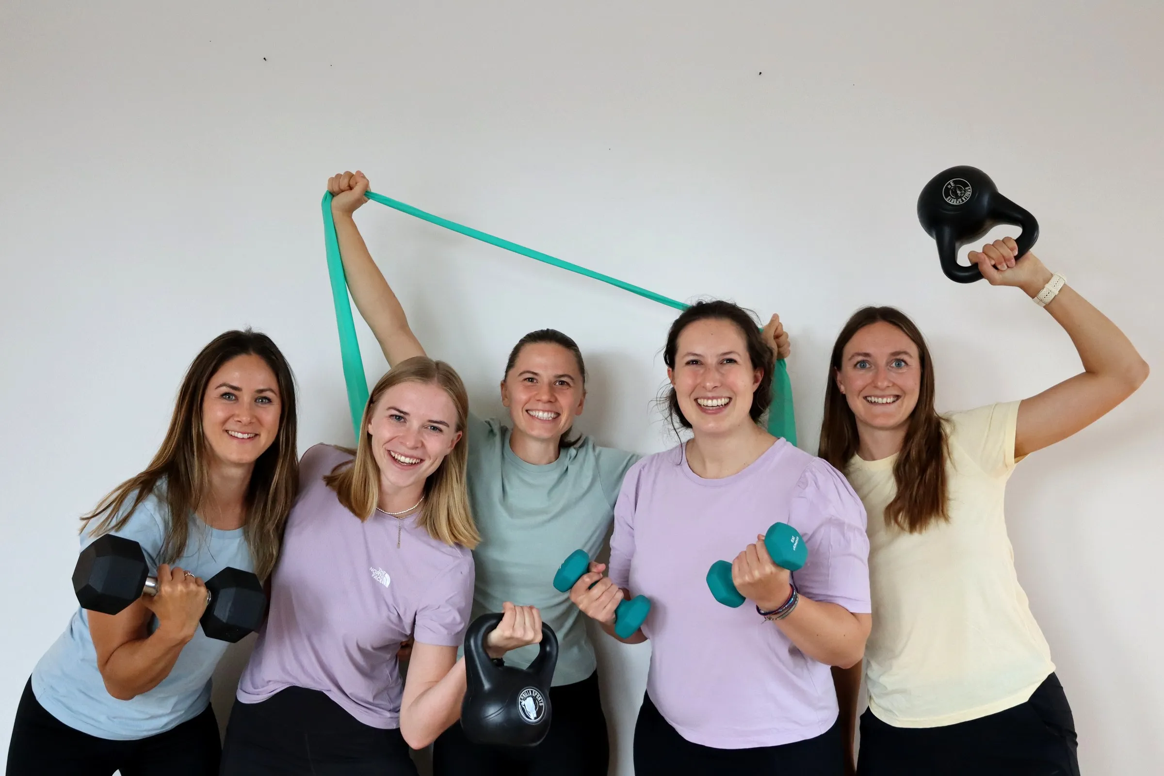 Die fünf Physiotherapeutinnen von doc+ Physio lachen in die Kamera, mit Kurzhanteln, Kettlebells und einem Trainingsband